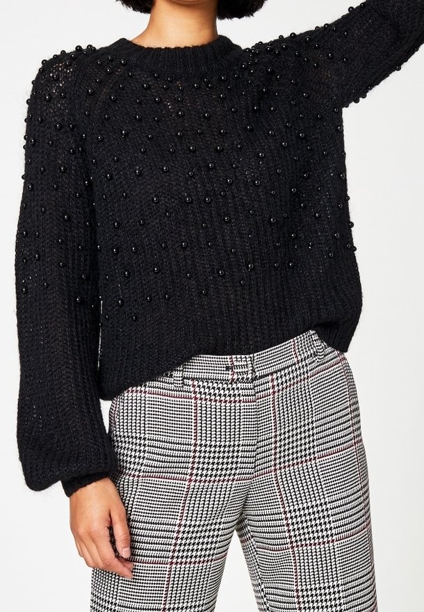 Pull noir en maille avec des accents de perles noires en relief, col rond, et coupe ample. Associé à un pantalon à carreaux taille haute avec des lignes rouges.