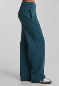Pantalones sudorosos de pierna ancha en color verde azulado, elaborados con tela suave, con una cintura acanalada, bolsillos laterales y una textura lisa. Combinados con zapatillas.