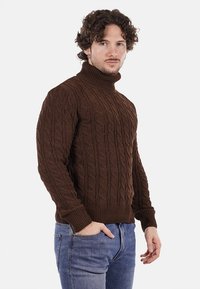 Maglione a collo alto in maglia marrone con un motivo testurizzato, caratterizzato da polsini e orlo a costine, indossato con jeans blu sbiaditi.