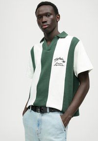 Groen en wit gestreepte polo met V-hals, korte mouwen en geborduurd logodetail op de borst, gecombineerd met lichtblauwe jeans.