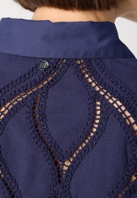 Gros plan sur le dos d'un vêtement bleu marine avec un motif en losange en dentelle ouverte et un petit bouton rond près du col, porté par une personne aux cheveux courts et bruns.