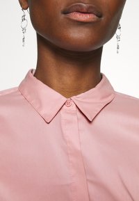 Rosa skjorta med klassisk krage, slät textur och en enkel knappstängning vid halsen. Silverörhängen med hängande detaljer.