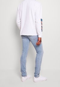 Witte, langdurige mouwen shirt met grafic op de linker mouw, gecombineerd met lichtblauwe skinny jeans en witte sneakers, gezien van de achterkant.