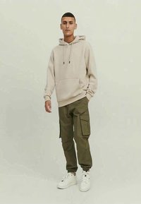 Jovem rapaz em pé, vestindo um hoodie bege, calças cargo verde-oliva e ténis brancos, contra um fundo claro e liso.