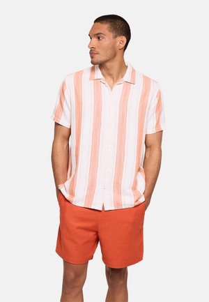 Hombre con camisa blanca de manga corta con rayas naranjas verticales y pantalones cortos naranjas lisos, de pie con las manos en los bolsillos, mirando hacia la izquierda.