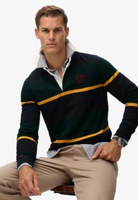 Polo-shirt met lange mouwen in groene, marineblauwe en gele strepen. Heeft een witte kraag en rode logodetail op de borst. Draagt met beige broek.