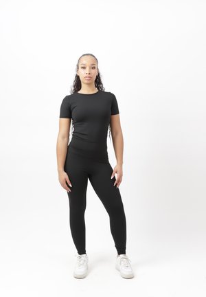 Kobieta stojąca przodem, ubrana w czarny dopasowany strój sportowy i białe buty sportowe na gładkim białym tle.