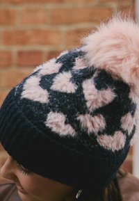 Close-up of a person wearing a black knit hat with pink fuzzy heart patterns and a large pink pom-pom against a brick wall background.