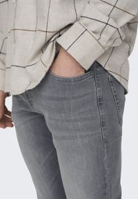 Jeanșii din denim gri au o textură netedă, fiind prevăzuți cu un design clasic cu cinci buzunare și un finisaj ușor decolorat. Sunt purtați cu o cămașă cu carouri deschise.
