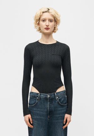 Ξανθιά γυναίκα που φοράει εφαρμοστή μαύρη μπλούζα με μακριά μανίκια και ψηλόμεσα άνετα μπλε τζιν, που στέκεται μπροστά σε ένα απλό λευκό φόντο.