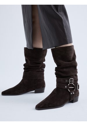 Botas holgadas de ante marrón oscuro hasta la mitad de la pantorrilla con punteras puntiagudas y correas con hebilla plateada, usadas con una falda de cuero negra.