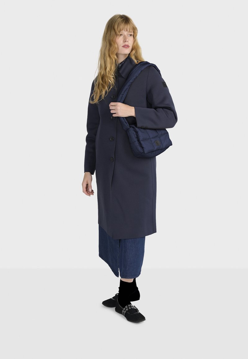 Cappotto lungo navy in tessuto elegante, con chiusura a bottone e cappuccio, abbinato a una borsa a spalla imbottita navy e culottes in denim.