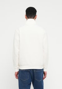 Felpa bianca con collo alto, polsini e orlo a coste. La parte posteriore presenta una superficie liscia, abbinata a jeans in denim blu.