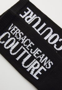 Fekete szövetpánt fehér hímzett szöveggel "VERSACE JEANS COUTURE" különböző méretekben és stílusokban, texturált kötött mintával.