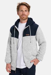 Sudadera con capucha y cremalleras, con un cuerpo gris y la parte superior azul marino, que presenta un tejido texturizado, cordones y un pequeño parche con el logo.