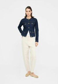 Denimová bunda v tmavě modré barvě s kulatým výstřihem, předním zapínáním na knoflíky, dvěma náprsními kapsami a knoflíky ve zlatém tónu, spárovaná se světle krémovými kalhotami.