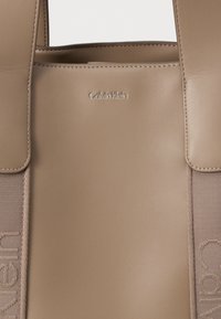 Calvin Klein beige læder tote taske med en glat tekstur, præget logo og en kontrasterende stofrem med trykt tekst.