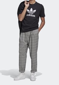 Černé bavlněné tričko s bílým logem Adidas, šedé kostkované kalhoty s puky a bílé tenisky, stojící na jednobarevném pozadí.