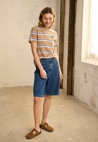 Femme debout à l'intérieur portant un t-shirt beige à rayures, un short en jean mi-long et des chaussures marron sans lacets, souriante et regardant vers le bas.