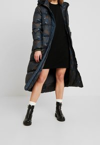 Longo cappotto imbottito blu scuro con motivo mimetico, con zip frontale, cappuccio e polsini a coste. Indossato sopra un vestito nero e stivali neri.