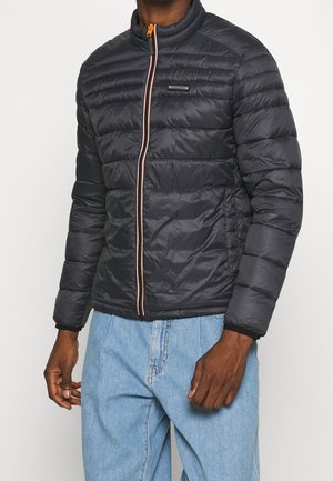 Mężczyzna w czarnej pikowanej kurtce z pomarańczowym suwakiem i jasnoniebieskich dżinsach, stojący na białym tle.