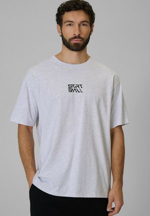 Man met baard, die een lichtgrijs T-shirt draagt met zwarte gestileerde tekst "START BALL", staand tegen een effen grijze achtergrond.