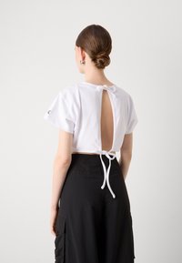 Λευκό cropped t-shirt με κοντά μανίκια και άνοιγμα στην πλάτη, ασφαλισμένο με έναν κόμπο. Φορεμένο με ψηλόμεσα μαύρα cargo παντελόνια που διαθέτουν πλαϊνές τσέπες.