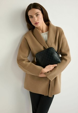 Kobieta o prostych brązowych włosach ubrana w camelowy płaszcz, trzymająca czarną pikowaną skórzaną torebkę na tle jasnego jednolitego tła.