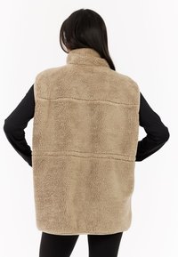 Gilet in pile color tan con colletto alto, texture morbida e dettaglio cucito sulla schiena. Indossato sopra una maglietta a maniche lunghe nera.