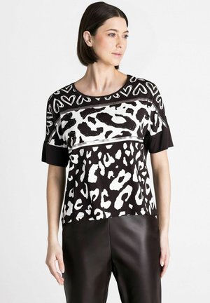 Vrouw die een zwart-wit T-shirt met korte mouwen draagt met hart- en dierenprints, gecombineerd met een zwarte broek, staand tegen een effen achtergrond.