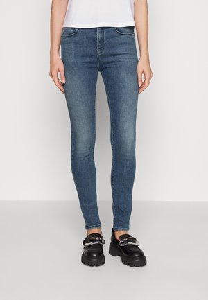 Woman wearing blue skinny jeans paired with black chunky loafers with silver buckle details against a plain background.