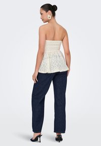 Kremfarbiges Peplum-Top mit einem gerafften Oberteil und Lochmuster, kombiniert mit dunkelblauen weiten Jeans und Absätzen, von hinten betrachtet.