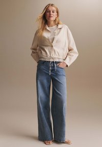Blonde woman stands with hands in pockets wearing beige buttoned jacket, beige knit top, wide-leg blue jeans, and beige open-toe sandals.