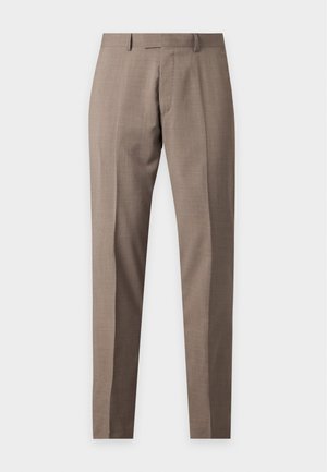 Homme debout portant une chemise bordeaux à manches longues boutonnée, un pantalon beige et des chaussures habillées noires sur un fond blanc uni.
