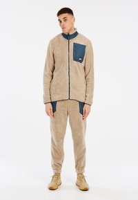 Giacca in pile beige con colletto alto e zip frontale, dotata di una tasca sul petto blu. Abbinata a pantaloni in pile beige e calzature color sabbia.