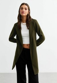 Olivgrön lång kavaj med skräddarsydd passform, slag och två sidofickor, bärs över en vit crop top och svarta byxor.