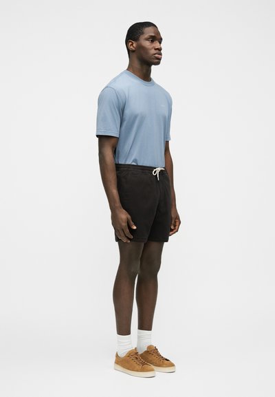 Jeune homme debout de profil portant un t-shirt bleu clair, un short noir à cordon de serrage, des chaussettes blanches et des baskets marron sur un fond uni.