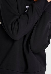 Sudadera negra con cuello redondo acanalado y puños; presenta una textura suave y un logo sutil en gris en la parte delantera.