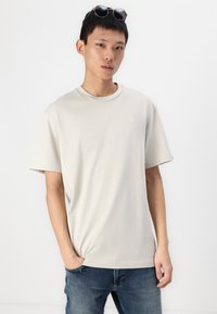 Lys beige bomuld T-shirt med rund halsudskæring, korte ærmer og et lille broderet logo på brystet. Båret med blå jeans.