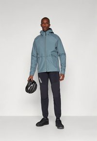 Vodoodporna modra jakna z kapuco, z zadrgo spredaj po celotni dolžini in nastavljivim spodnjim robom. Opremljena s črnimi oblazinjenimi hlačami in črno čelado za kolo.