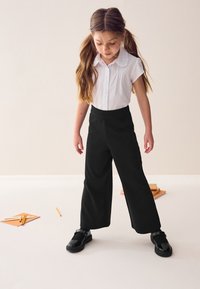 Mustat leveälahkeiset housut pehmeästä kankaasta, yhdistettynä valkoiseen lyhythihaiseen kauluspaitaan ja mustiin kiiltonahkaisiin kenkiin, joissa on hihna.