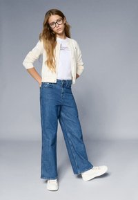 Bela kardigan brez rokavov, pletena tekstura nad belo srajco z vijoličnim napisom. Modri široki jeans in bele superge. Preprost, sproščen outfit.