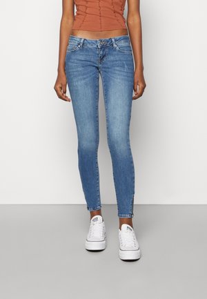 Jean bleu skinny avec une taille mi-haute, cinq poches et des détails d'ourlet effiloché. Associé à des baskets blanches et un haut texturé.