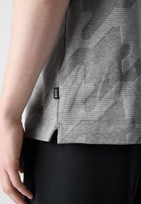 Gros plan sur le côté d'une personne portant une chemise grise à motifs avec une petite étiquette "BOSS" et un pantalon noir visible.