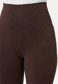 Gros plan sur une personne portant des leggings taille haute de couleur marron foncé avec un petit logo "LES LUNES" brodé près de la hanche.