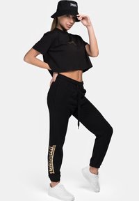 Black cropped t-shirt, black joggers with gold graphic text, paired with white sneakers and a black bucket hat. Smooth fabric and relaxed fit.