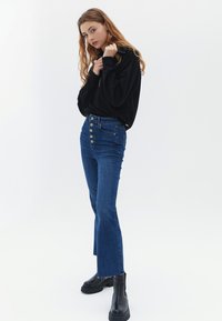 Jeans flare biru denim dengan pinggang tinggi, dilengkapi dengan penutup lima tombol dan ujung kain yang mentah. Atasan lengan panjang hitam dengan detail ritsleting.