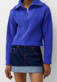 Pull en maille bleu vif avec un col large et une demi-fermeture éclair, associé à une mini-jupe en denim foncé avec un ourlet roulé.