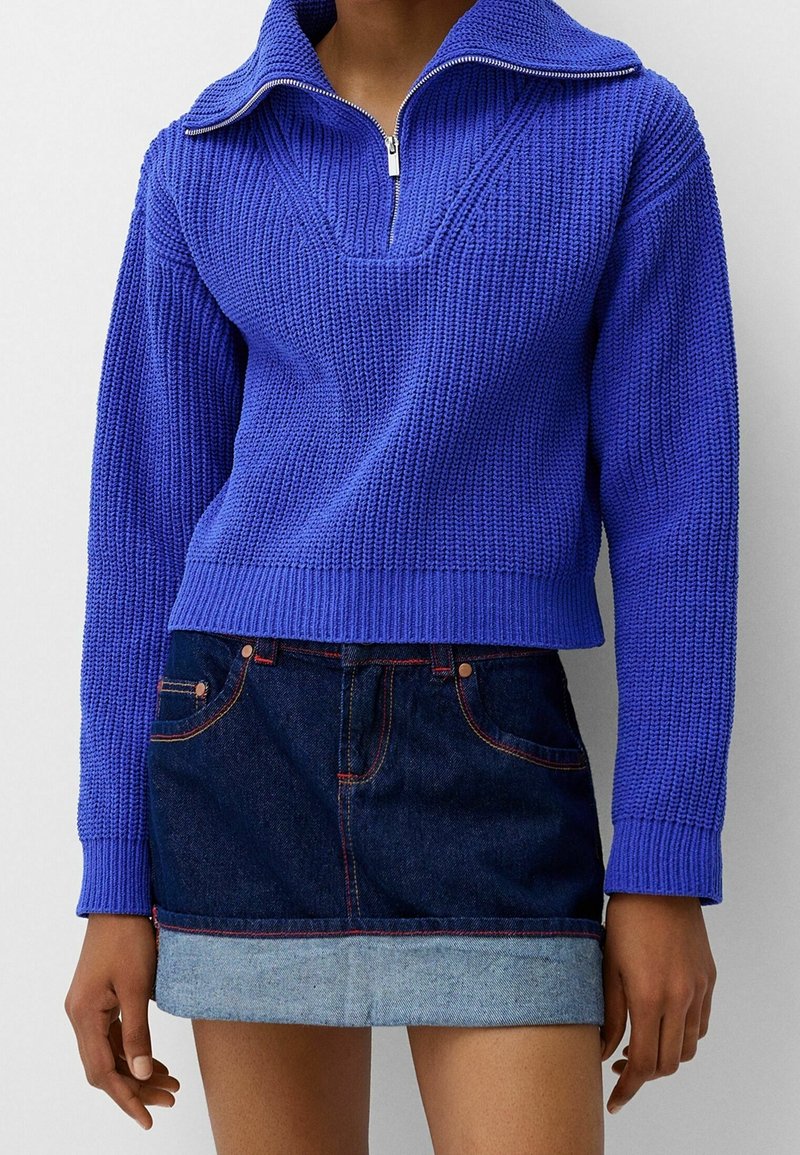 Pull en maille bleu vif avec un col large et une demi-fermeture éclair, associé à une mini-jupe en denim foncé avec un ourlet roulé.