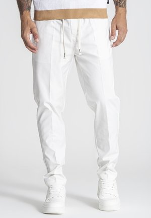 Muž oblečený v bielych nohaviciach so sťahovacou šnúrkou a bielych teniskách, stojaci s uvoľnenými rukami pozdĺž tela na jednoduchom pozadí.